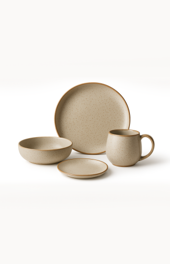 A minimalist stoneware dinnerware set in a speckled beige finish with a natural brown rim is arranged against a plain white background. The collection features a large dinner plate, a medium bowl, a small saucer, and a matching mug.