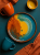 A top-down view on a vibrant ceramic plate with an abstract orange and teal glaze centered on a dark teal background. The artistic composition includes a matching teal mug, an orange mug, a small yellow bowl, and a crumpled terracotta fabric.