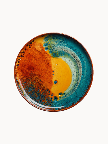 A top-down view on a flat ceramic plate with a vibrant, artistic glaze with a fiery orange center bleeding into a deep teal border.