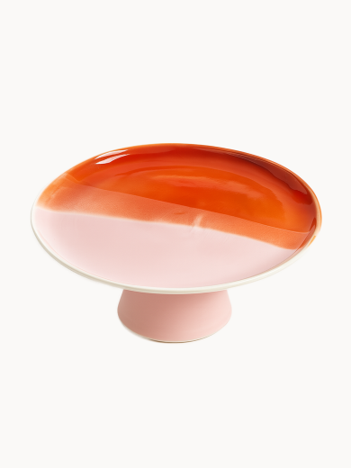 A ceramic cake stand with a two-tone surface, on a white background. The smooth top is half glazed in glossy amber-orange and half in matte pastel pink, supported by a short pink pedestal base.